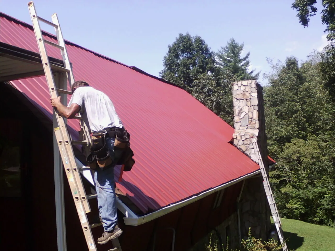 Roofer on a ladder beside a red metal roof and stone chimney — Soffit & Fascia Repair by Roofing Brothers in Menasha